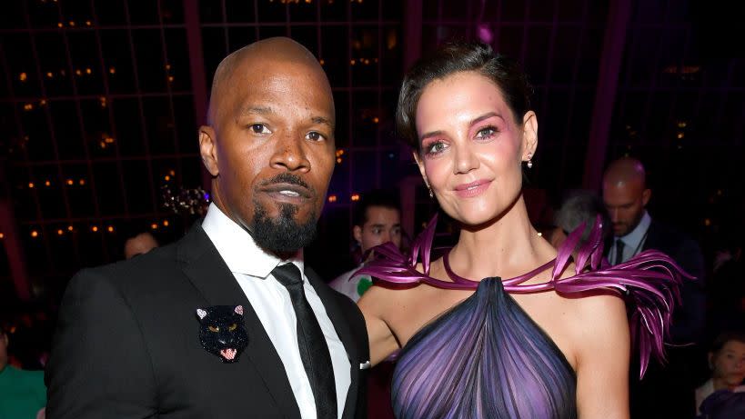 jamie foxx and katie holmes stand and pose for a photo, he wears a black suit with a tie and a sparkly panther pin, she wears a purple gown and makeup