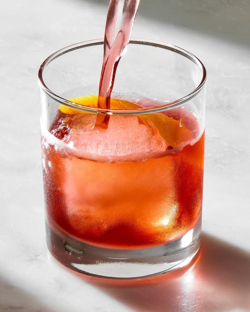 angled shot of a negroni being poured over a large ice cube in a small rocks glass.
