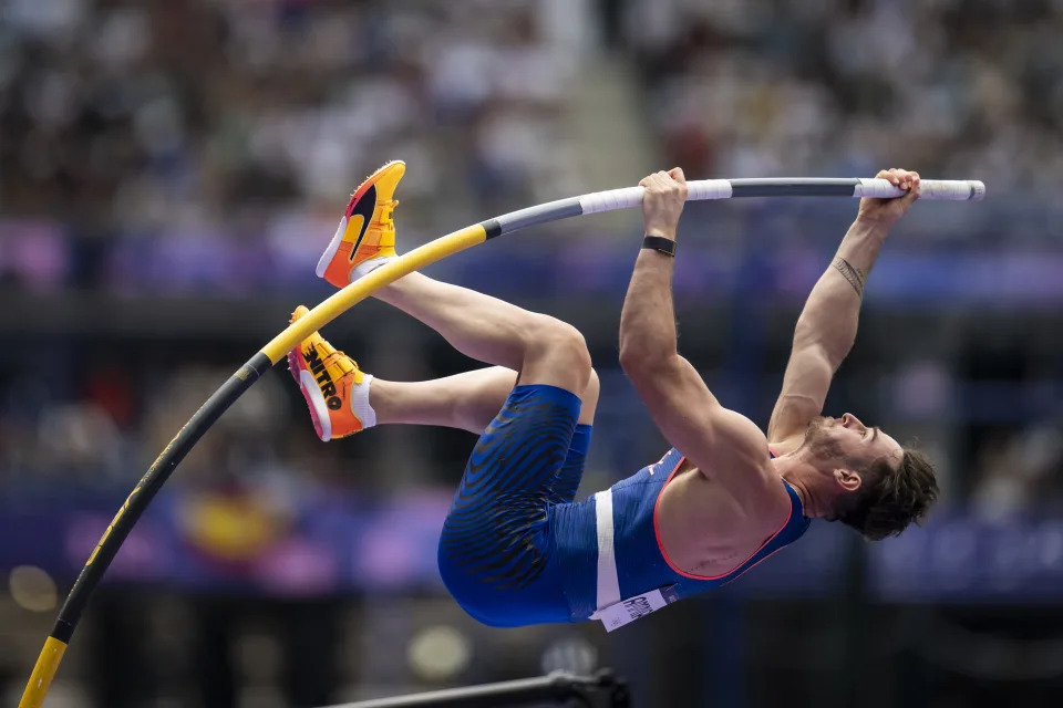 A pole vaulter, unidentified and in mid-air, performing at a track and field event. The background is a blurred crowd