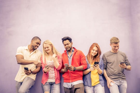 Five adults leaning on a wall with smartphones in their hands.