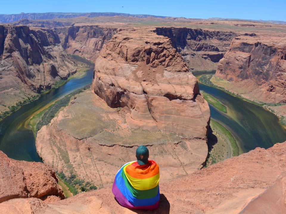 A person wrapped in a pride flag sits in front of small red canyon with water running through it and blue skies in the background.