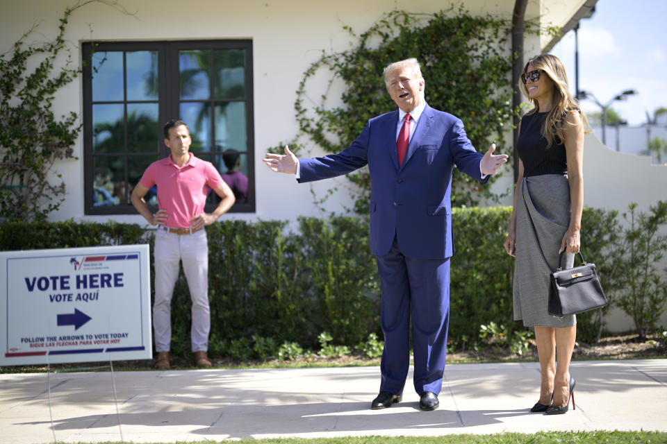 Former President Donald Trump spreads his arms expansively as his wife stands by his side, looking cheerful.