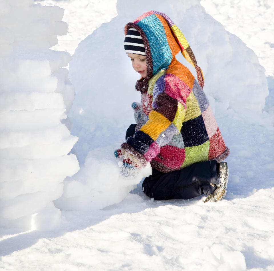 toddler wearing colourful patchwork coat, snow pants, and striped toque playing outside in white snow, snow fort