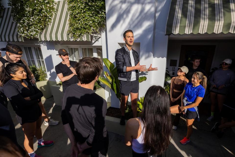 A man addresses a crowd of runners outside a building with striped awnings.