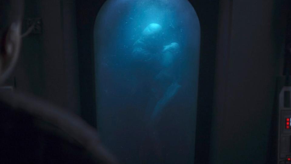 Greef Karga looks at a small Snoke-like body floating in blue liquid in a vat on The Mandalorian