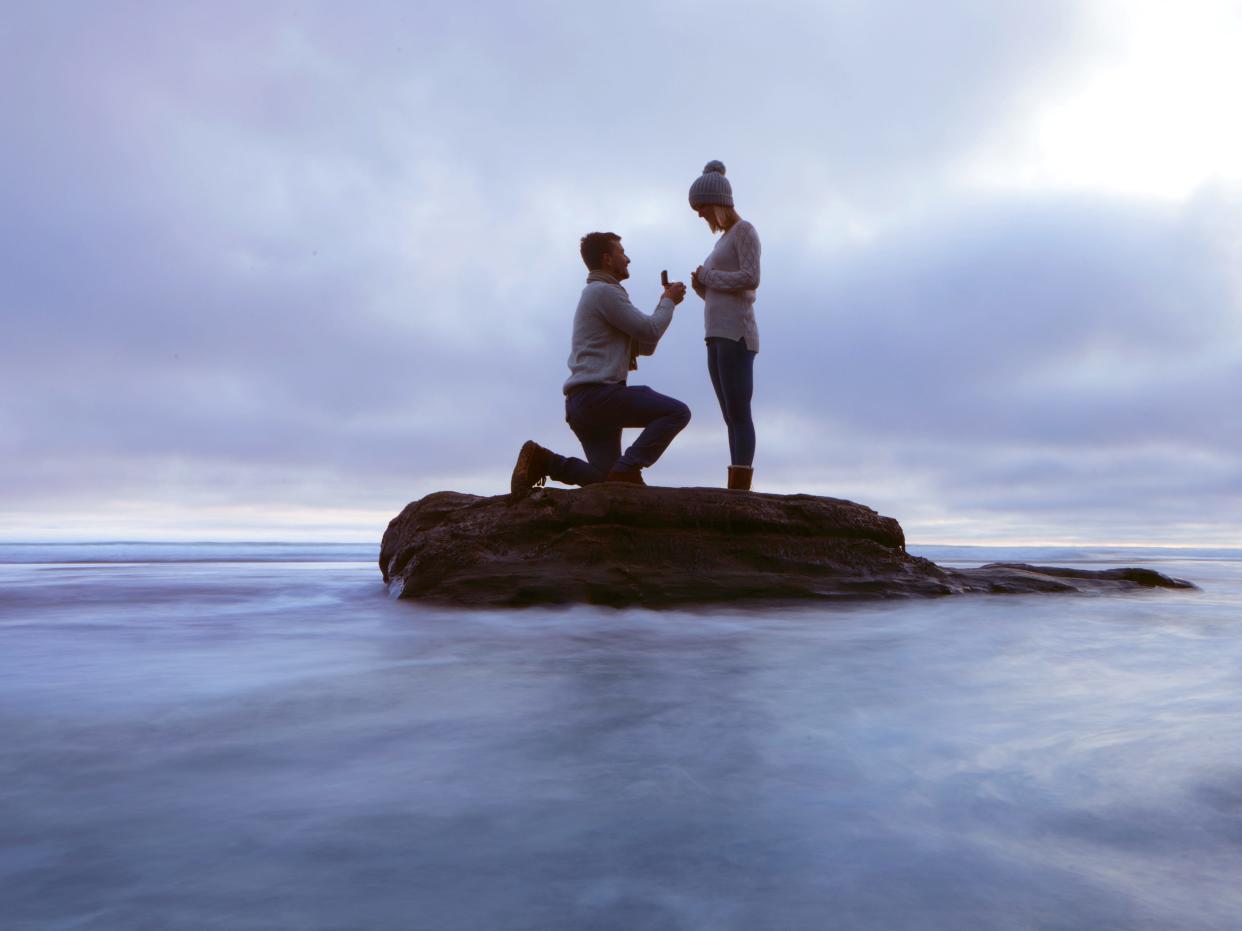 A stock image of a man proposing to a woman on a rock on a lake.