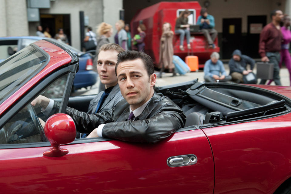 Two gangsters with slick-back hair drive along a futuristic street in a convertible