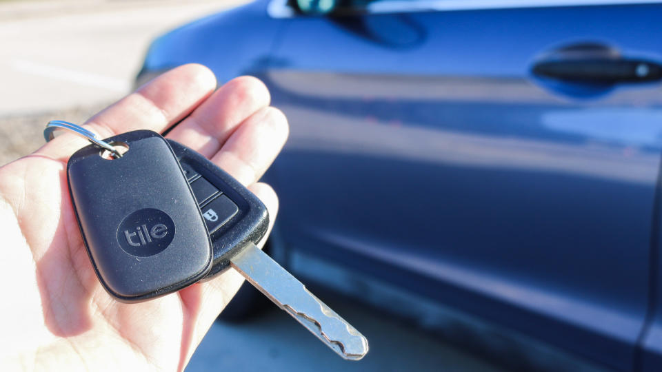 A hand holding a pair of keys with a Tile Pro tracker in front of a car