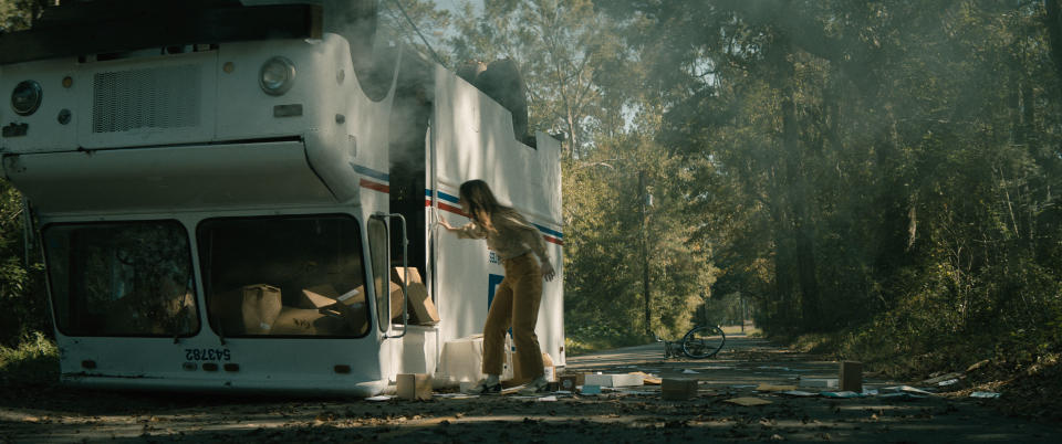 a young woman on a wooded street peers inside a turned-over panel truck