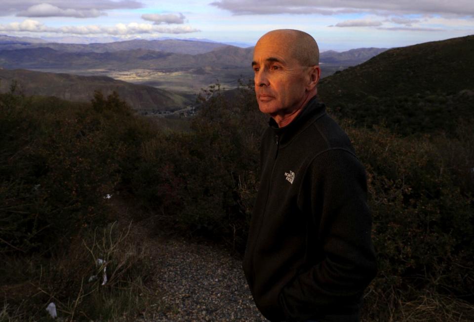 A man with very close-cropped hair stands outdoors with mountains in the distance.