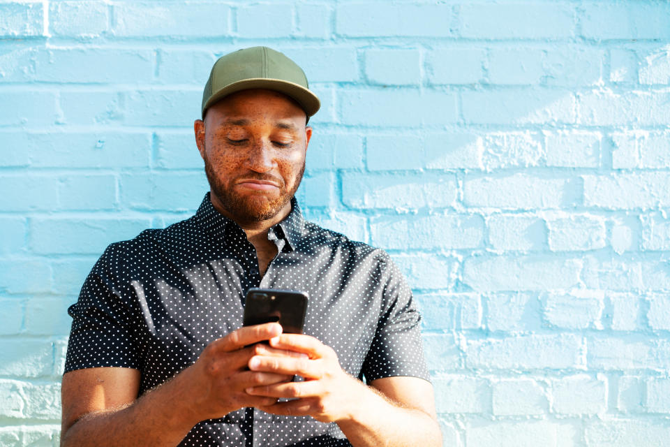 Man in a cap and dotted shirt using a smartphone against a blue brick wall