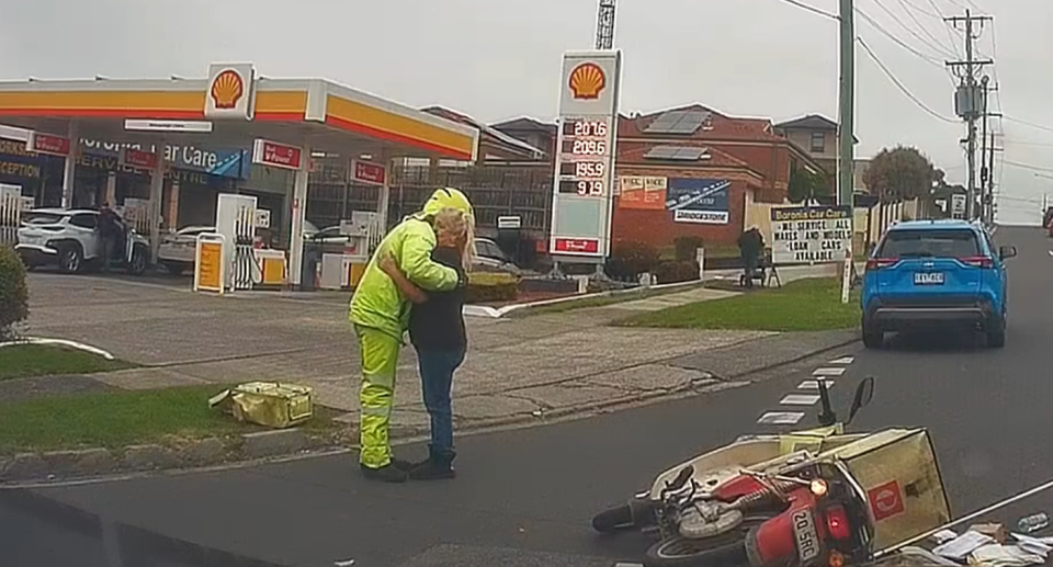 The postie wearing hi-vis hugs the woman who came from the blue car as the Australia Post bike lies on its side on the road. 
