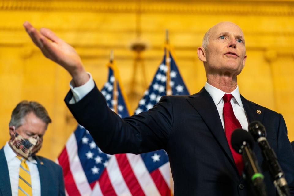 Rick Scott in a suit and red tie speaking into microphones while gesturing with his right arm raised.