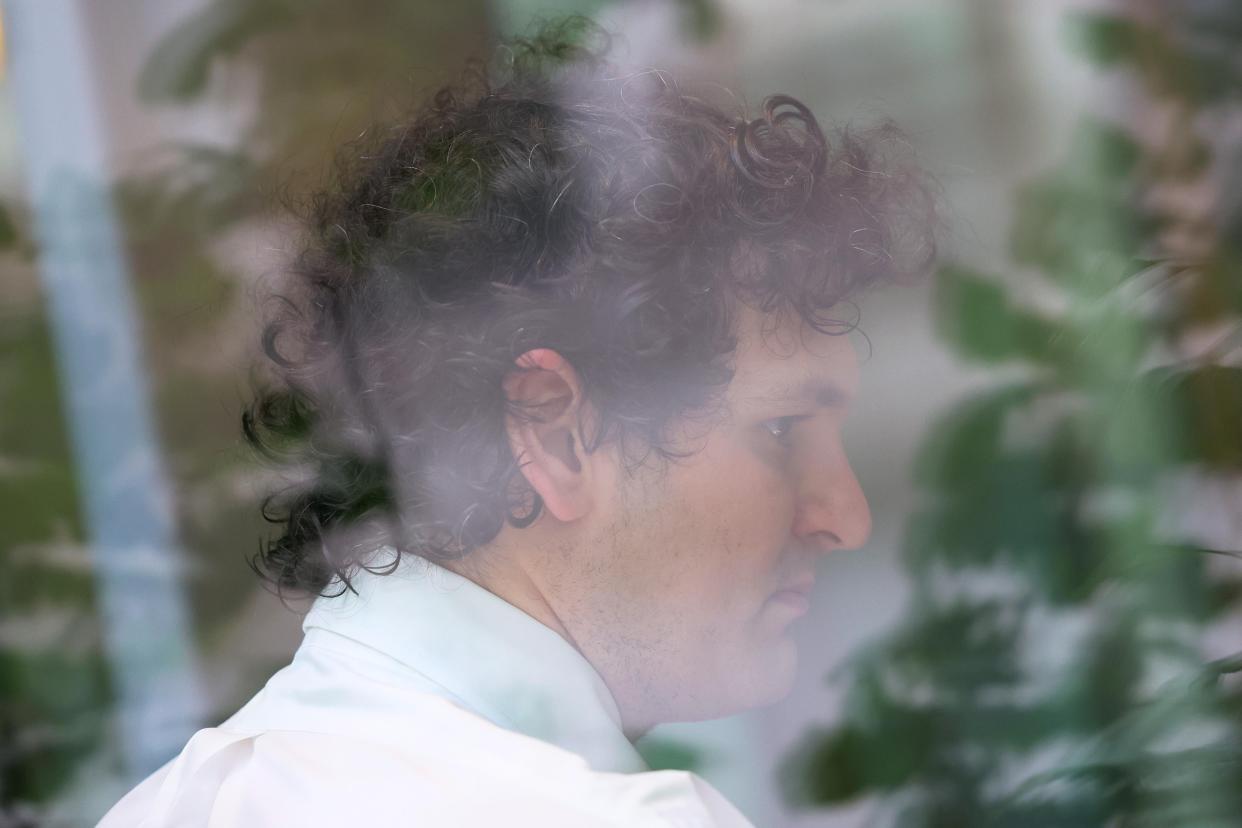 Through a window, the side of a man's face. He has curly dark hair and is wearing a white button-down shirt.
