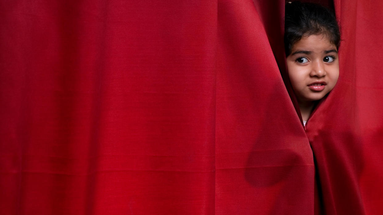  Little girl peeking out from a red stage curtain with a nervous expression. 