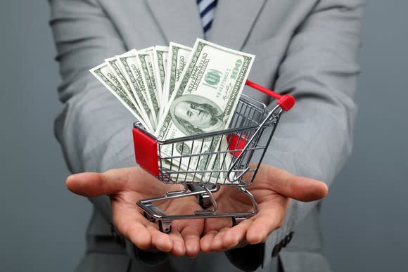 A man in a suit holds a tiny shopping cart full of money.