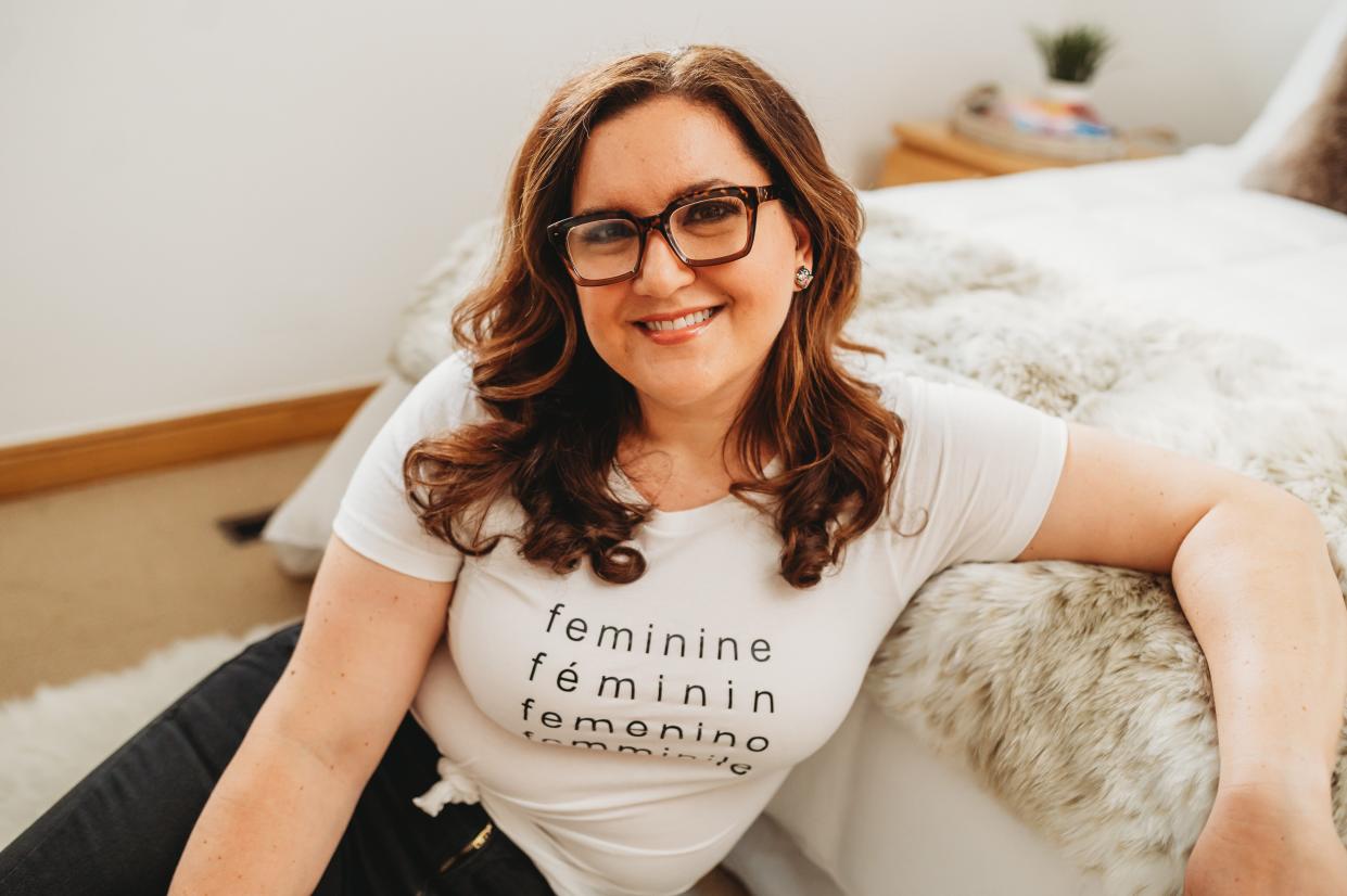 Andrea Javor wearing glasses, a white t-shirt, and jeans sitting on the ground and smiling on the camera while leaning on a bed.