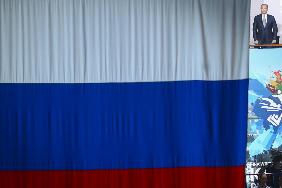 FILE - In this July 24, 2015 file photo Russian President Vladimir Putin is seen at right next to the Russian national flag as he listens to the anthem of Russia during the opening ceremony at the Swimming World Championships in Kazan, Russia. The World Anti-Doping Agency banned Russia on Monday Dec. 9, 2019 from the Olympics and other major sporting events for four years, though many athletes will likely be allowed to compete as neutral athletes. (AP Photo/Denis Tyrin, file)