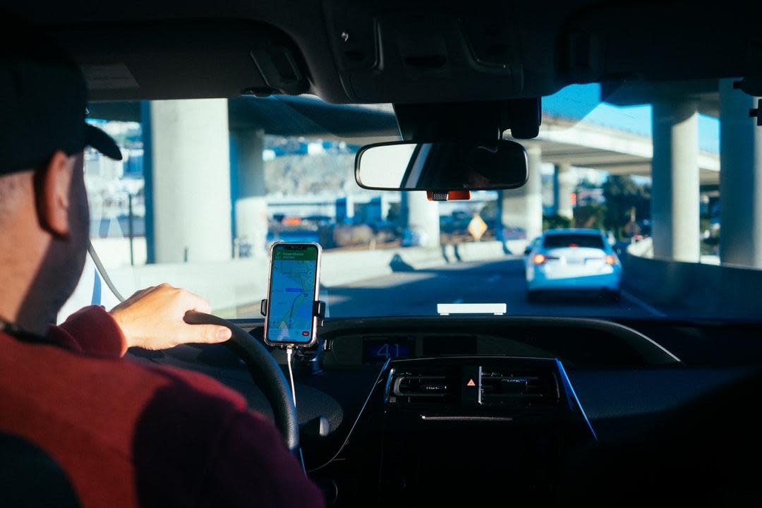 A view from the back seat of a Uber driver with a baseball cap driving down a highway using his smartphone map app. (Stock Image)