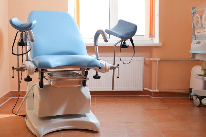 Gynecological examination chair in a medical room with a window and medical equipment in the background