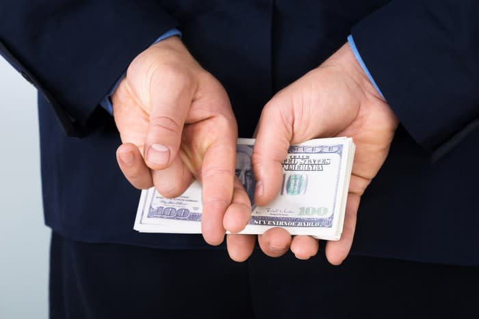 A businessman holding a stack of cash behind his back, with his fingers crossed.