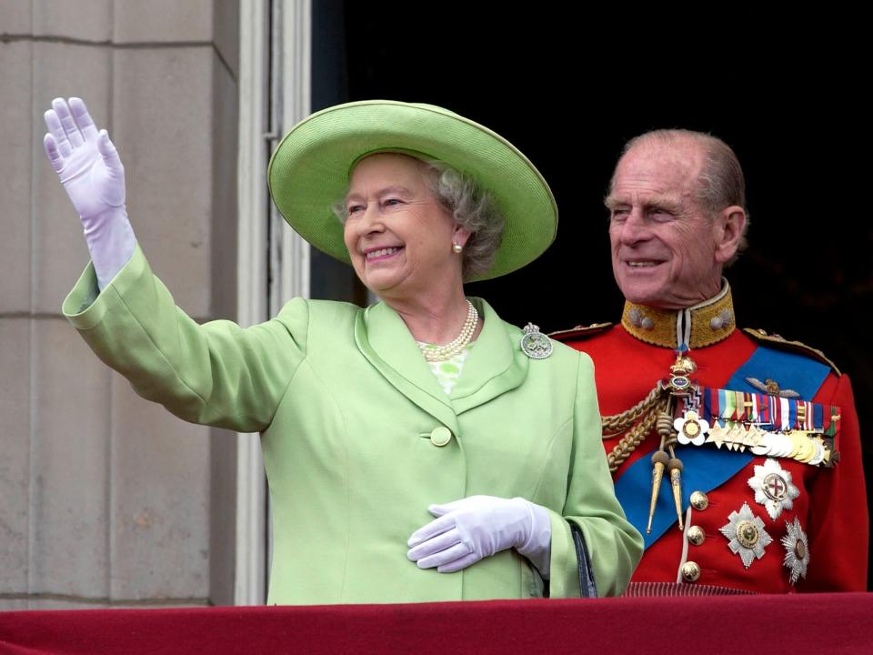 The Queen smiling and waving in a muted lime green overcoat with round satin collars and large buttons. You can slightly see a white dress with a green circular pattern. She's wearing a matching green wide-brimmed hat with a necklace of pearls and white gloves.