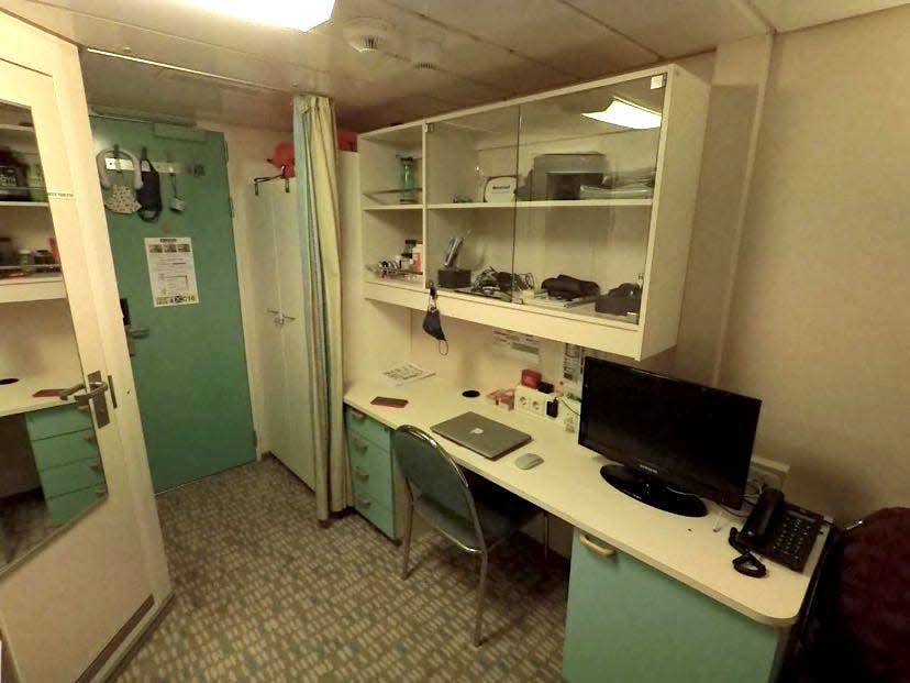 Crew cabin on cruise ship that is white and blue with a door, desk, and cabinet