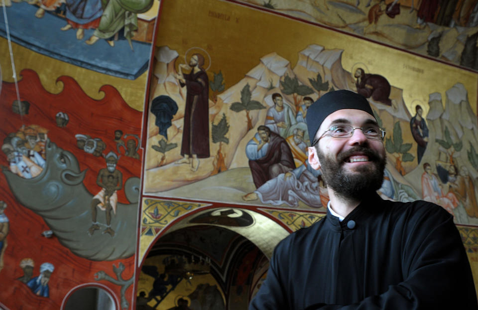 In this photo taken Tuesday, Feb. 4, 2014 Orthodox priest Branko Vujacic smiles in front of the brightly-colored newly painted fresco, left, in the Serbian Orthodox Church of Christ's Resurrection in Montenegro's capital Podgorica. The fresco allegedly shows late Yugoslav autocratic leader Josip Broz Tito drowning in red fiery waves of hell with Karl Marx and Friedrich Engels, authors of the 1848 Communist Manifesto. They are in the company of Adam and Eve, together with some of Montenegro's current politicians and people wearing Muslim turbans. What appear to be rival church priests are being swallowed through a huge jaw of an angry gray beast with pointed devil ears. The fresco has triggered much attention and public controversy in this tiny former communist country. (AP Photo/Risto Bozovic)