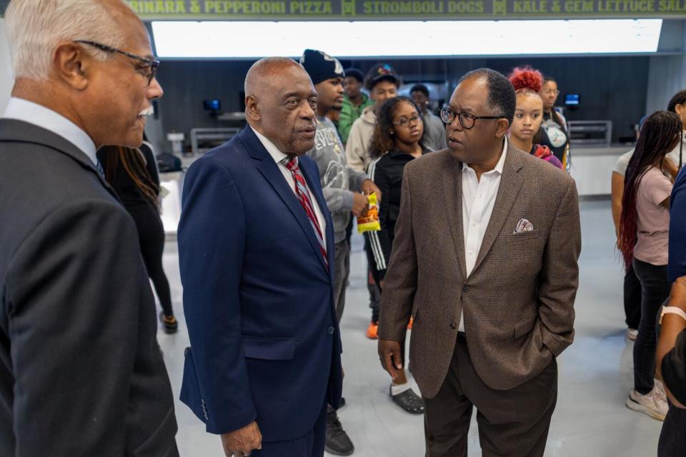 Three men standing and talking in a large room as other people mill around behind them