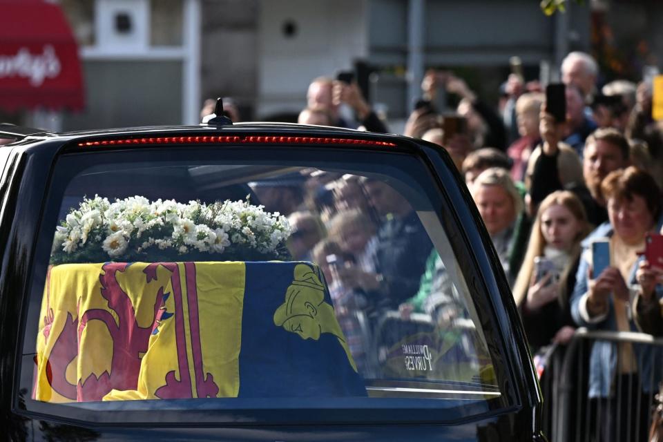 TOPSHOT - Members of the public pay their respects as they hearse carrying the coffin of Queen Elizabeth II, draped in the Royal Standard of Scotland, is driven through Ballater, on September 11, 2022. - Queen Elizabeth II's coffin will travel by road through Scottish towns and villages on Sunday as it begins its final journey from her beloved Scottish retreat of Balmoral. The Queen, who died on September 8, will be taken to the Palace of Holyroodhouse before lying at rest in St Giles' Cathedral, before travelling onwards to London for her funeral. (Photo by Paul ELLIS / AFP) (Photo by PAUL ELLIS/AFP via Getty Images)