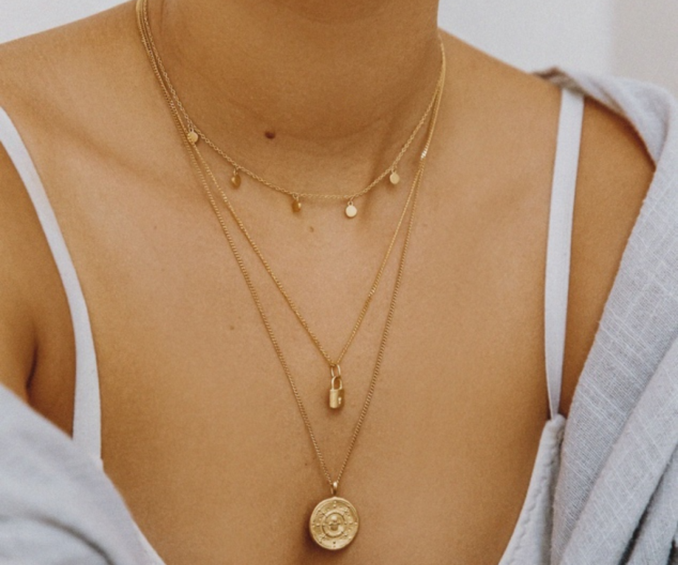 An olive skinned woman wears layers of fine gold necklace chains against a pale grey background.