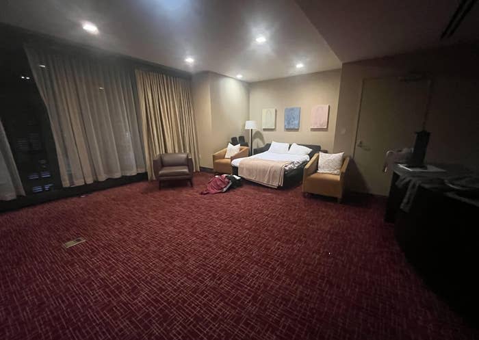 A hotel room with dim lighting, featuring a bed, nightstands, a sofa, an armchair, a desk, and several bags and clothes on the floor