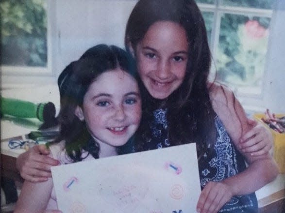 Lena Crown and her friend Talya as kids. The girl on the left has dark brown hair, and freckles and wears a light, sleeveless top. The girl on the right is taller and wears a navy sleeveless top with a white pattern. Together, they hold up a piece of paper with drawings on it.