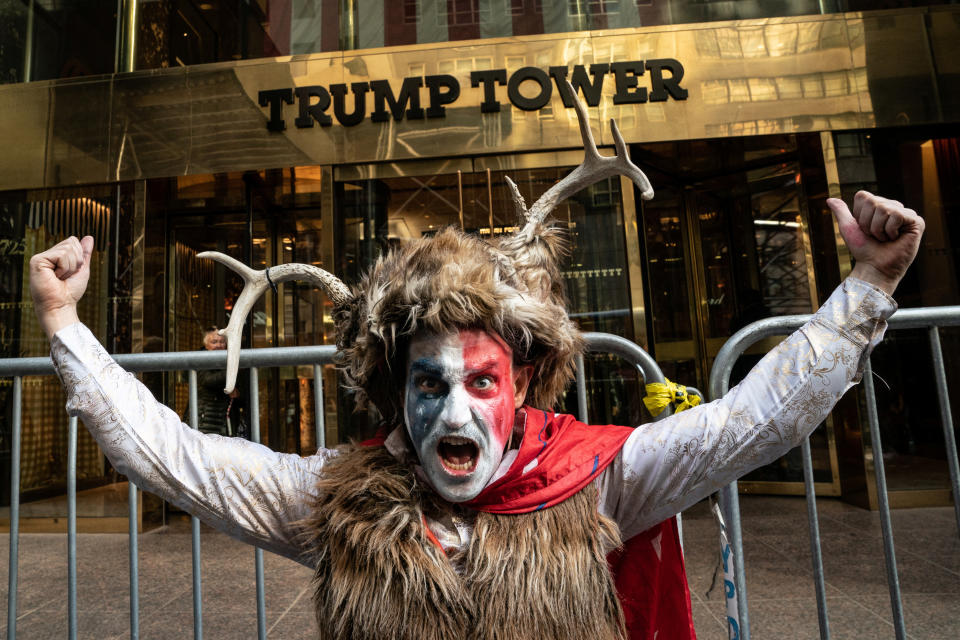 A demonstrator with red, white and blue face paint wearing a fur vest and a fur hat decorated with two antlers, raises his fists in the air and shouts in front of the entrance to Trump Tower.