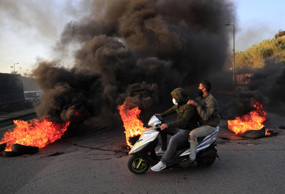 Protesters ride a scooter past burning tires set on fire to block a highway that leads to Beirut's international airport, in Beirut, Lebanon, Tuesday, March 2, 2021. Scattered protests broke out in different parts of Lebanon Tuesday after the Lebanese pound hit a record low against the dollar on the black market, a sign of the country's multiple crises deepening with no prospects for a new Cabinet in the near future. (AP Photo/Hussein Malla)