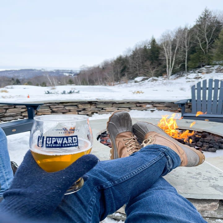 A snowy scene in the distance and a person wearing jeans and holding a beer in a glass labeled Upward Brewing Company in the foreground near a fire pit