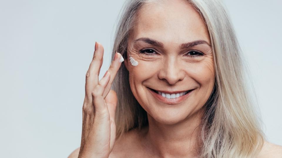 A woman with grey hair applying a cream to her face to get rid of rosacea permanently