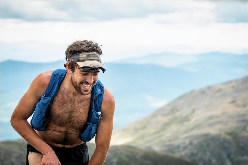 A man wearing a running vest and visor tops a hill with mountains in the background, Jack Kuenzle FKT Presidential Traverse