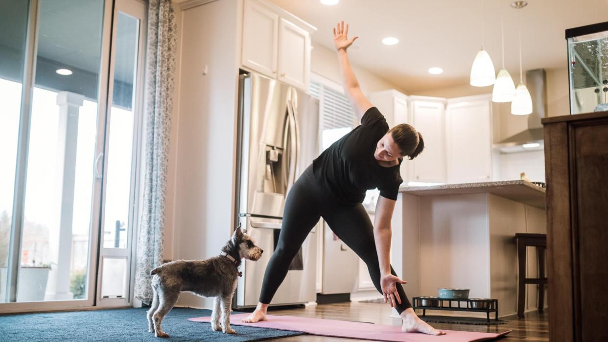 A mature adult woman does yoga and strength training exercises on a mat in her living room, her pet terrier dog keeping her company and trying to play.