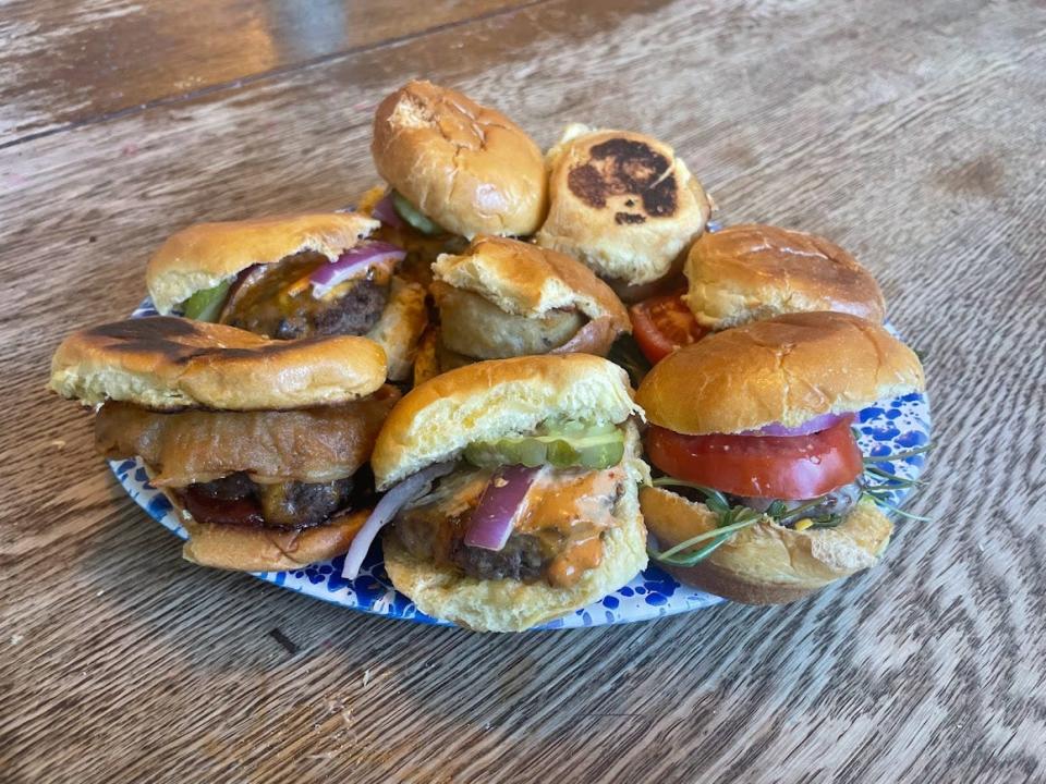 A blue paper plate on a wooden table filled with sliders. Some sliders have toasted buns, a tomato slice, pickles, onion rings, and raw onions