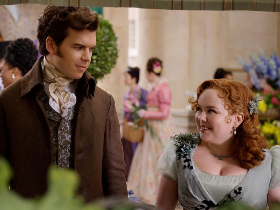 colin bridgerton and penelope featherington, played by luke newton and nicola coughlan, standing together and smiling at each other in a floral market