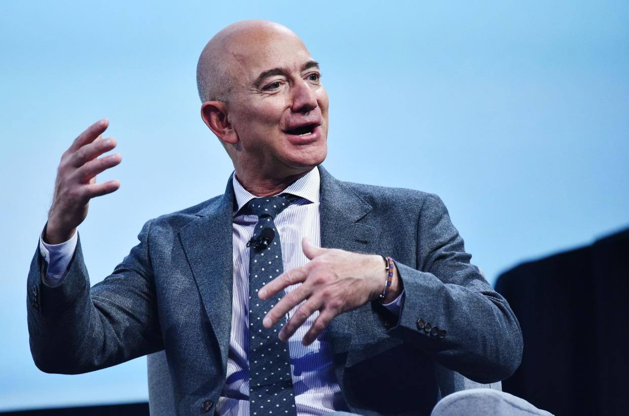 Amazon's former CEO Jeff Bezos smiles and gestures with his hands while talking in front of a pale blue background.