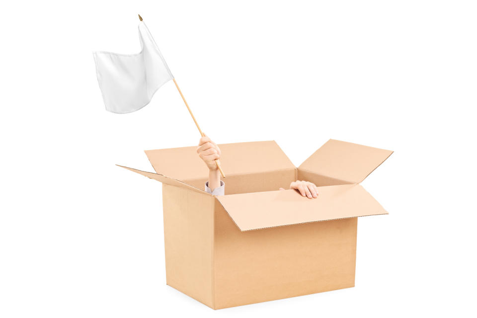 A man hiding in a cardboard box waves a white flag.