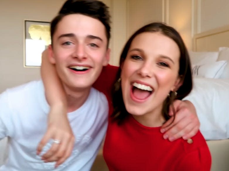 noah schnapp and millie bobby brown with their arms around each other, smiling and sitting in front of a bed in a hotel room