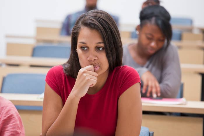 Woman in a red top sitting at a desk in a classroom with another person behind her. She appears thoughtful