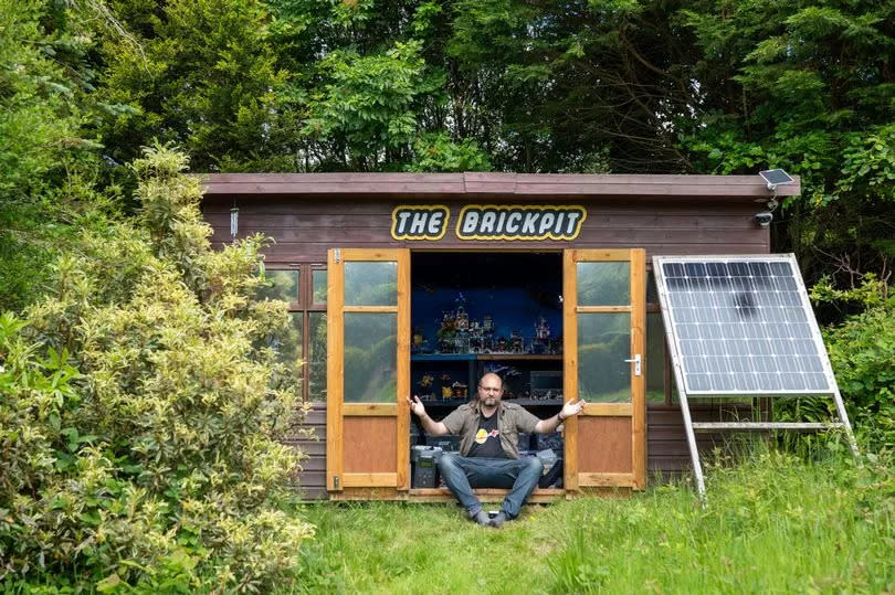 A man wearing jeans sat in the doorway of a cabin adorned with a sign that reads 'The Brickpit'