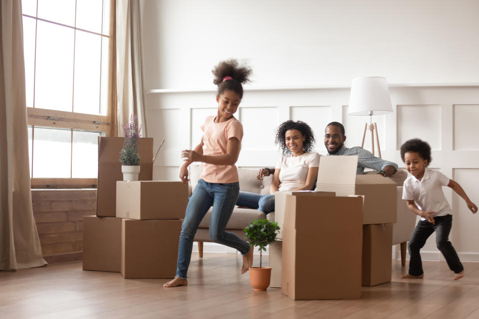 Active funny kids play run around cardboard boxes having fun on moving day with black parents sit on sofa, happy african american family children mom dad laughing in living room celebrate relocation