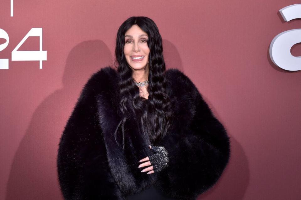 Cher poses on the red carpet in a luxurious black fur coat and sparkly gloves, smiling for the camera