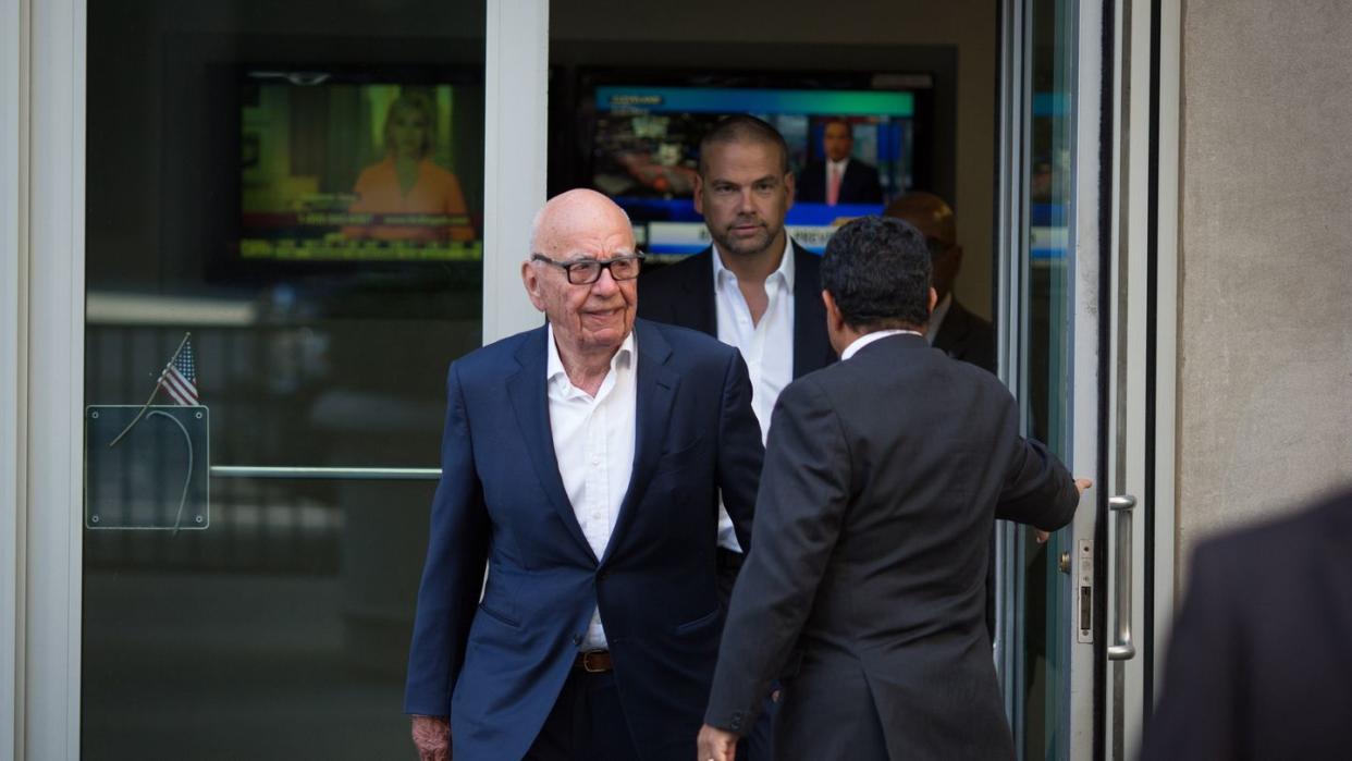 rupert murdoch and lachlan murdoch walk out a set of glasses doors that a man holds open, all three men wear dark suits and white collared shirts