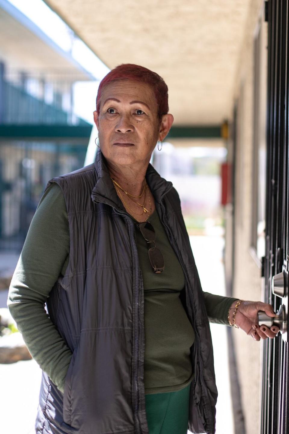 A woman stands outside with her left hand on a door handle.